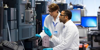 Two technicians confer working on an LC-MS equipment bank in a lab.