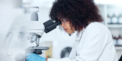 One mixed race scientist with curly hair wearing safety goggles and gloves analyzing medical test samples on a microscope in a lab. 