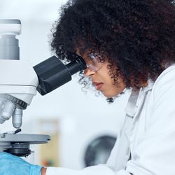 One mixed race scientist with curly hair wearing safety goggles and gloves analyzing medical test samples on a microscope in a lab. 