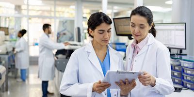 Two female scientists in lab wearing lab coats looking at a clip board