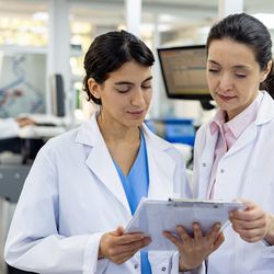 Two female scientists in lab wearing lab coats looking at a clip board