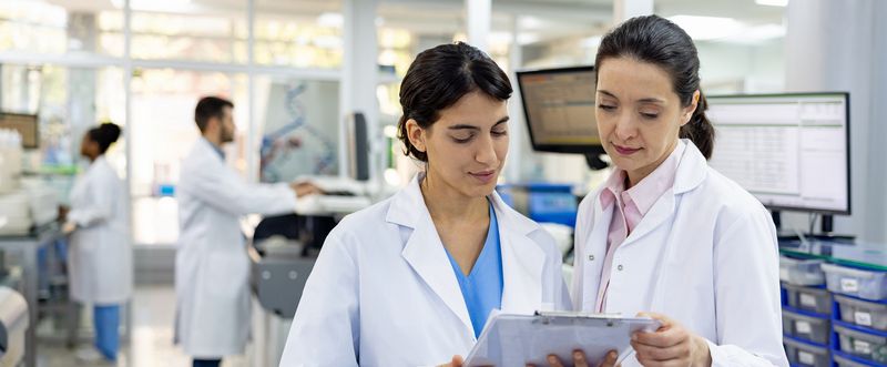 Two female scientists in lab wearing lab coats looking at clip board