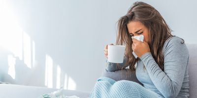 Sick woman sits on the couch holding a cup of tea and sneezing into a tissue