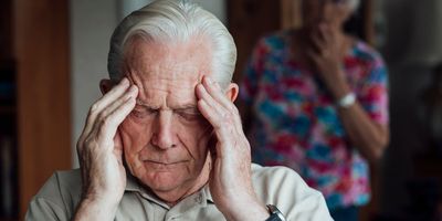 Elderly man rubs his temples while his wife looks on with concern