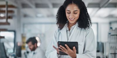 Female researcher in white lab coat and dark curly hair using a tablet in a lab setting