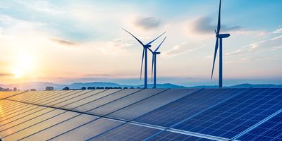 A photo of an expanse of solar panels and three wind turbines behind them and a city skyline and hills in the background