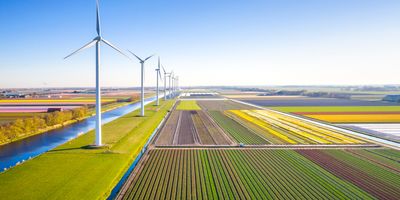 an aerial view of a row of wind turbines next to a neatly cultivated colorful farm field with a canal on their other side