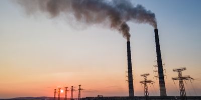 a pair of smokestacks emitting plumes of smoke on top of a factory with powerlines behind it as the sun sets in the background