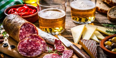 sliced salami on a wooden cutting board, with sliced cheese, a bowl of olives, a bowl of chili peppers, and two glasses of beer surrounding it, all on a wooden table