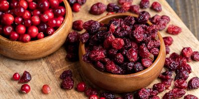 two wooden bowls, one containing fresh and one containing dried cranberries sit on a wooden table surrounded by other loose cranberries