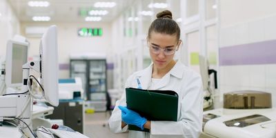 A female scientist, with her brown hair in a bun and wearing safety glasses, a lab coat, and blue gloves, makes notes on a green clipboard while standing next to various machinery in a lab.