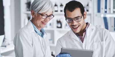 two scientists, an older woman with short grey hair and a younger man with short dark hair and stubble, smile broadly as they discuss something they are looking at on a tablet. Both scientists are wearing white lab coats, glasses, and blue gloves.