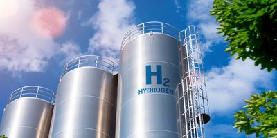 a low angle view of three very large silver cylindrical hydrogen storage tanks against a backdrop blue sky with fluffy white clouds and green tree leaves on the right edge of the image