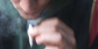 close up of a man wearing a dark green sweater holding a lit rolled cigarette or joint in his hand, showing only the lower half of his face, slightly out of focus and obscured by smoke