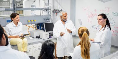 a group of male and female scientists having a discussion in a lab with a whiteboard in the background