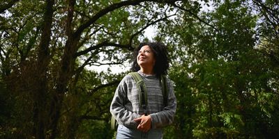 a woman with brown skin and dark curly hair in a gray sweatshirt looks happily at the trees around her while on a walk outdoors