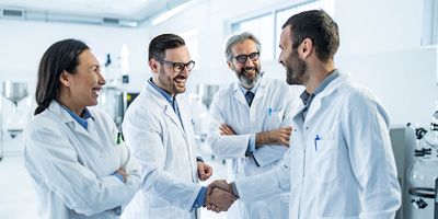 Four scientists in lab coats are standing in the lab in a group smiling. Two of the men are shaking hands.