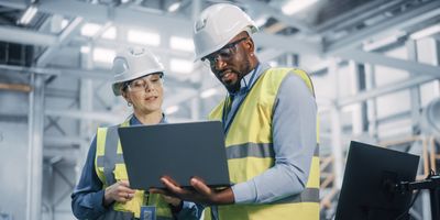 Two safety inspectors in hard hats and safety vests use a laptop in a laboratory