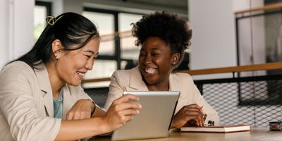 two cheerful young businesswomen sitting at table and laughing while watching a video on digital tablet.