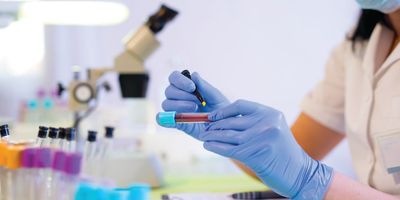 Female scientist wearing gloves holds a testable filled with red liquid in her left hand and prepares to label the test-tube with a marker in her right hand.  