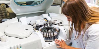 A female lab professional in white lab coat opens a piece of equipment for maintenance.