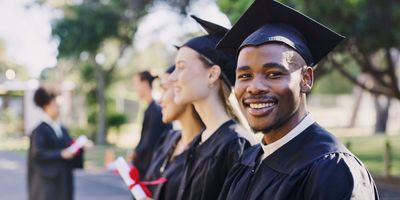 smiling graduates in black gowns and graduation caps, the black man is looking directly at the camera with two white woman standing beside him and looking off camera, one is holding a diploma tied with a red ribbon, all are standing on a tree lined path