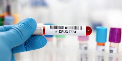 a blue-gloved hand holds a blood sample vial that is labeled "drug test" with other blood sample vials visible in the background