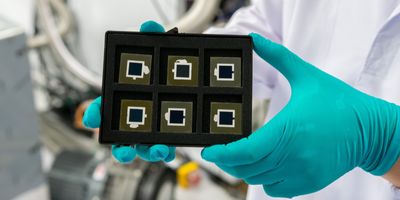 hands in aqua gloves hold a box containing six black and white squares that are the new solar cells made of silicon and perovskite, with lab tech in the background