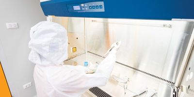 A fully gowned, gloved, and hooded lab worker pipettes a liquid at a biological safety cabinet in the lab.