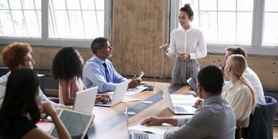 A young, new leader stands at the head of the boardroom table. She is wearing a white blouse and grey skirt and is pointing as she discusses important details with her staff., who are all seated at the table.