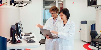 A male and female scientist in lab coats discuss something on the female scientist's computer while standing in the lab.