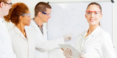 A young female scientist stands with a clipboard while three of her colleagues work on a whiteboard in the background. Everyone is wearing lab coats.