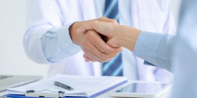 A closeup of a handshake between a lab-coated scientist and a businessperson in a dress shirt over top of a desk with a clipboard and tablet to illustrate the choosing of a CRO.