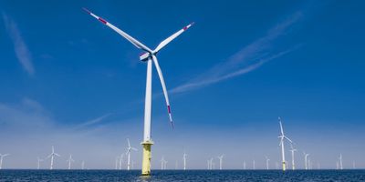 one close large white wind turbine in an offshore windfarm with many other wind turbines and a mostly clear blue sky in the background