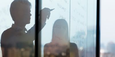 Two people in silhouette working on a glass board