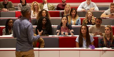 A professor, facing away from the camera, teaching a class of undergrads, facing the camera