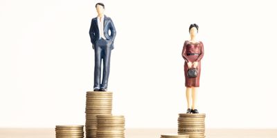 Male and female figurines on uneven piles of coin representing the pay gap on a white background