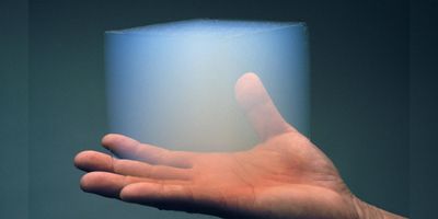 a hand holding a smoky translucent cube of aerogel, on a dark teal background