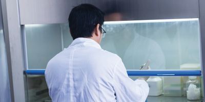 An image of a male researcher wearing a white lab coat and gloves sitting at a fume hood.