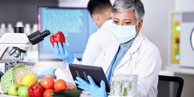 An older female scientist wearing a blue mask and gloves and a white lab coat holds a red pepper while looking at a tablet. Many other fruits and vegetables are sitting on a lab bench in front of her next to a microscope, while a man works on a computer in the background