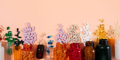 a photo of multiple pill bottles and containers laying flat in a line on a peach colored surface with various shapes, sizes, and styles of pills spilling out of them