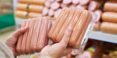 two packages of hotdogs or sausages being held in front of a shelf of other processed meats in a store