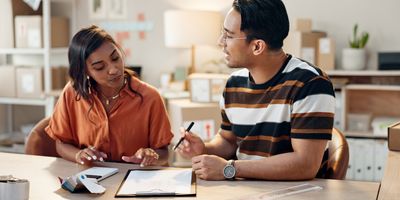 a man with tan skin and dark hair wearing a brown striped shirt talks to a brown skinned woman with black hair in an orange shirt while she is looking at a clipboard on a desk, in an office setting