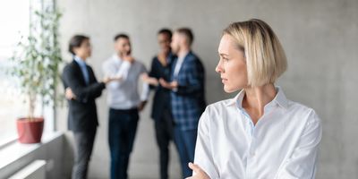 a blond business woman with a bob in a white button down shirt stands looking off to the left with her arms crossed while a group of business men in suits discuss something in the background