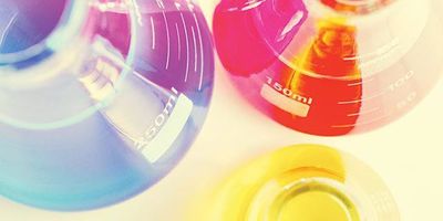 An overhead closeup image of three beakers on a lab bench. The largest beaker, which is on the left side, has blue liquid in it. The medium beaker, which is to the right of the large beaker, has red liquid, while the smallest beaker, at the front of the image, is filled with bright yellow liquid.