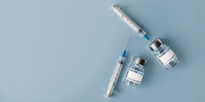 two glass vials with white labels that say "vaccine" and two white syringes with blue needle tips arranged next to each other on a sky blue surface