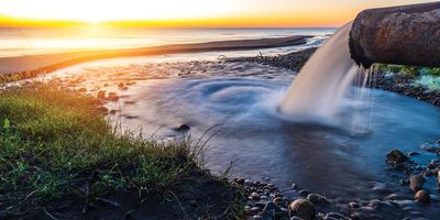 a rusty pipe with water pouring out of it into a large lake or ocean at sunset