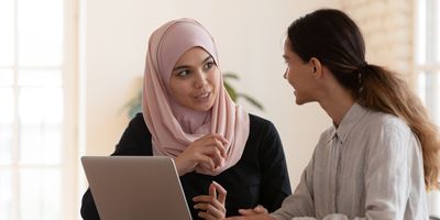 a woman in a pink hijab speaks to another woman with brown hair in front of a laptop