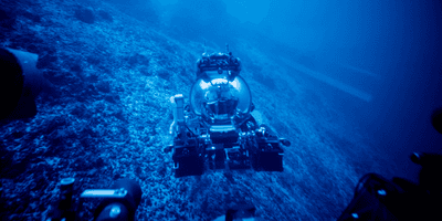 Two men in a bubble shaped submersible just above the ocean floor, everything is blue-hued. Chief Scientist Frieder Klein and Deep Rover Pilot Alan Scot exploring a submerged carbonate platform.