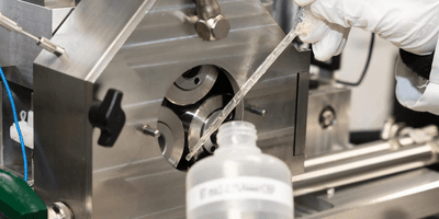 a white gloved hand uses a pipette to move a clear liquid lubricant from a plastic bottle to rotating parts of a large machine